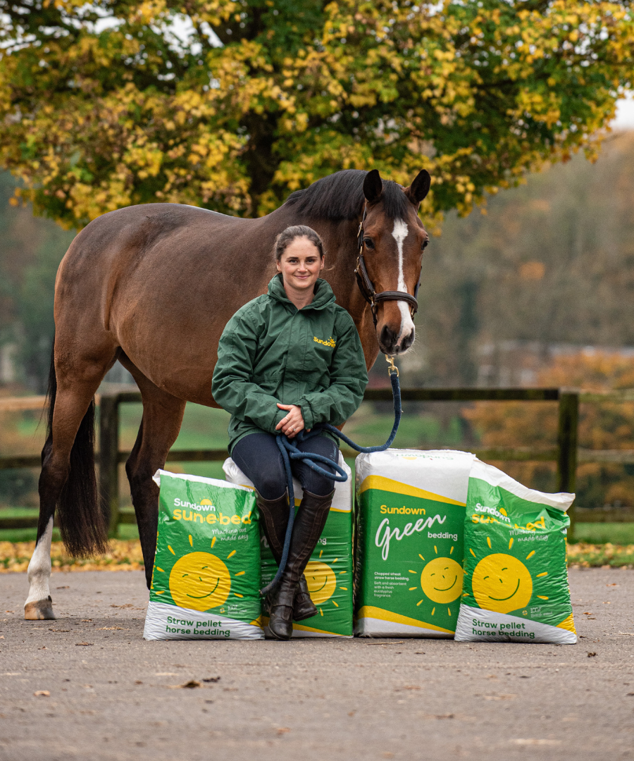 Woman sitting on bales of sundown horse bedding, with a brown horse standing behind her