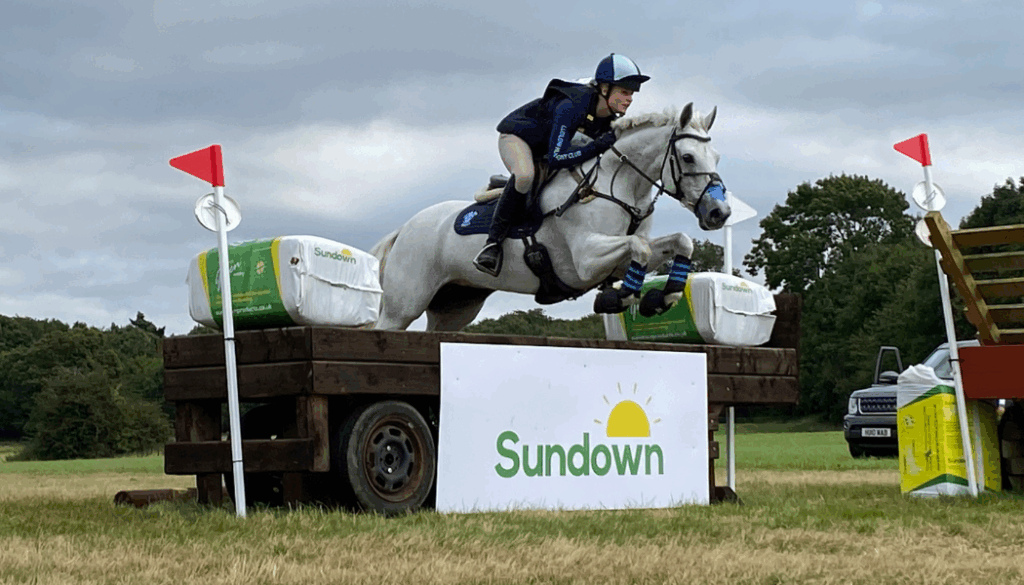 A grey horse jumps over a cross country fence with Sundown bedding bales